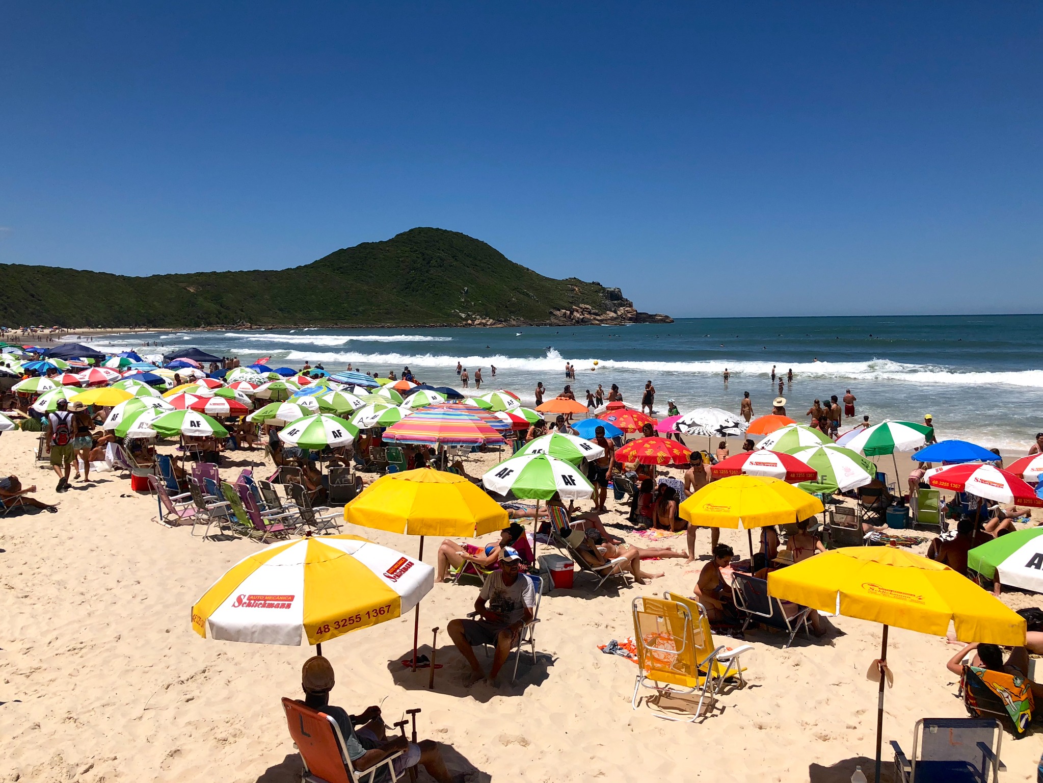 Praia movimentada durante o verão, com o mar e o morro ao fundo